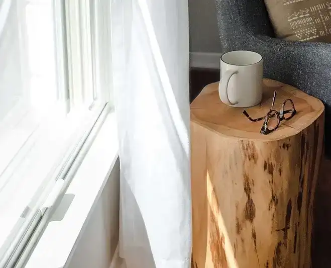 Bright light shines through a Marvin Replacement window with a coffee mug sitting on a wood log table.